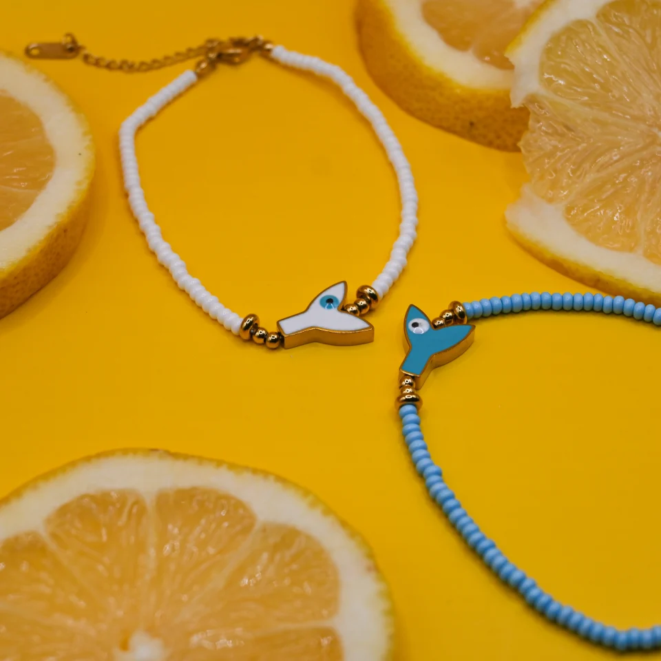 Summer mermaid tail white anklet with evil eye in stainless steel. Featuring a gold plated steel chain that fluctuates, white glass grommets and a central steel element with mermaid tail and peephole design. Water-resistant summer jewelry for every woman.