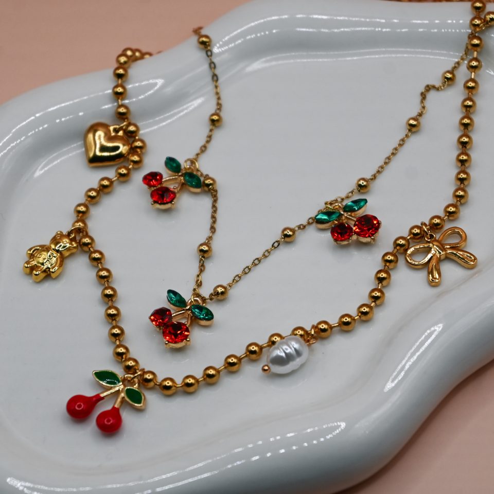 Gold steel charm necklace with cherries, teddy bear, pearl, heart, and bow steel charms on a trendy steel bead chain. Bold spring jewelry for 2026. Hot summer jewelry trends for 2026.