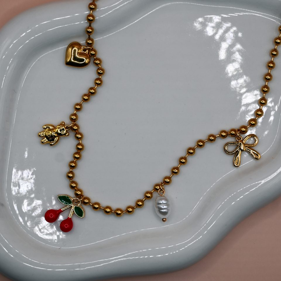 Gold steel charm necklace with cherries, teddy bear, pearl, heart, and bow steel charms on a trendy steel bead chain. Bold spring jewelry for 2026. Hot summer jewelry trends for 2026.