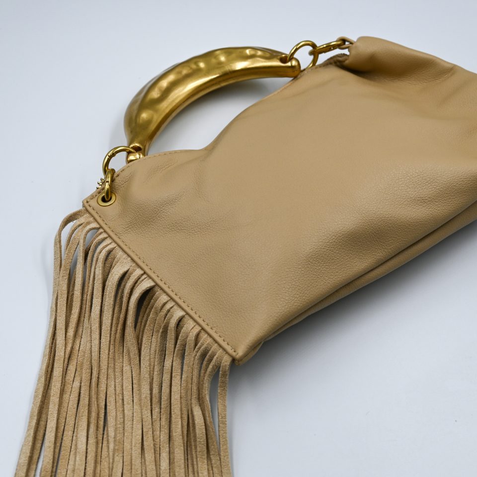 Gloria sand handbag, crafted from high-quality synthetic leather with a smooth texture. It features a statement metal handle in a gold shade with a hammered finish. It includes decorative suede tassels and a detachable leather strap.