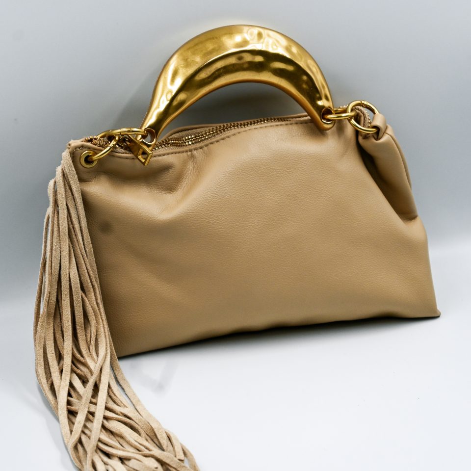 Gloria sand handbag, crafted from high-quality synthetic leather with a smooth texture. It features a statement metal handle in a gold shade with a hammered finish. It includes decorative suede tassels and a detachable leather strap.