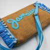 Natural raffia summer clutch featuring light blue decorative tassels, Jamaica embroidery, detachable fabric wrist strap, and adjustable crossbody strap.