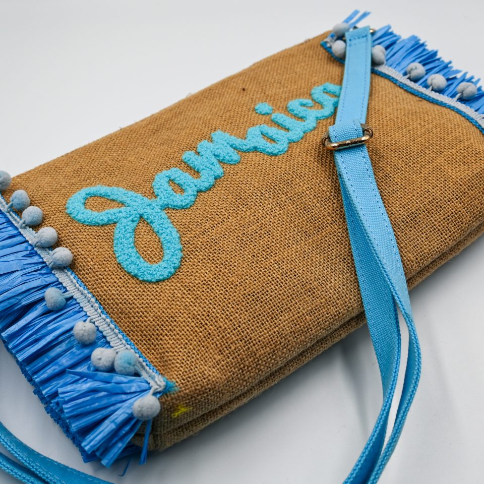 Natural raffia summer clutch featuring light blue decorative tassels, Jamaica embroidery, detachable fabric wrist strap, and adjustable crossbody strap.