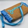 Spacious raffia clutch bag with embroidered Jamaica lettering, light blue tassels, zipper closure, detachable hand strap, and adjustable shoulder strap.