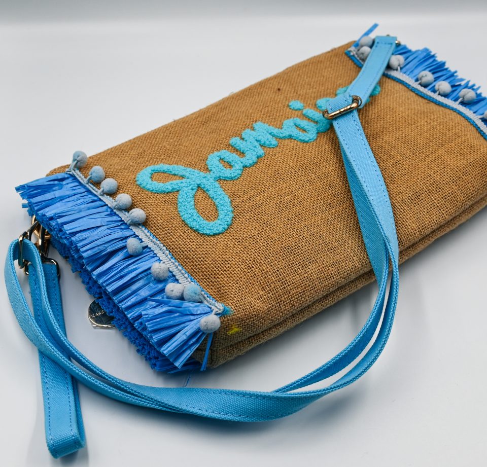 Spacious raffia clutch bag with embroidered Jamaica lettering, light blue tassels, zipper closure, detachable hand strap, and adjustable shoulder strap.